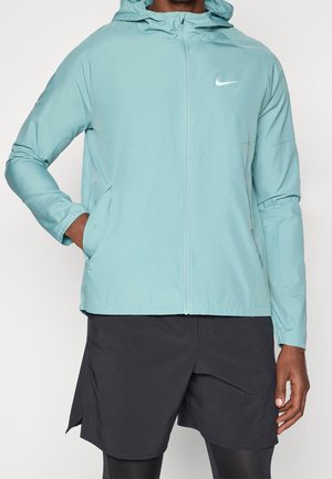 Mężczyzna w jasnoniebieskiej bluzie Nike z kapturem i zamkiem błyskawicznym, czarnych szortach i legginsach, stojący z jedną ręką w kieszeni.