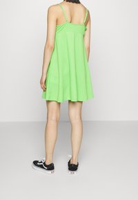 Grön bomullsklänning med spaghettistil, strukturerad tyg, utsvängd nederdel, bärs med svarta sneakers och vita snören, sedd bakifrån.