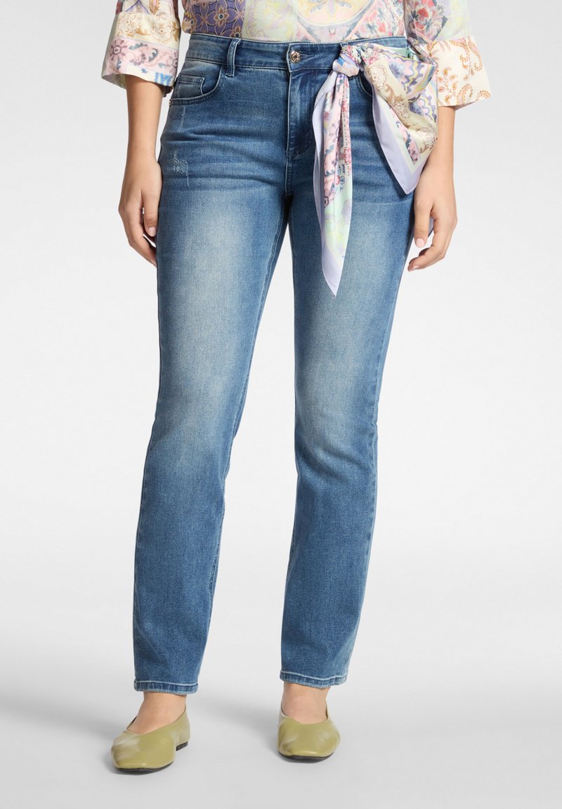 Jeans di denim blu con vestibilità a vita media, lieve scoloritura e una sciarpa chiara annodata in vita. Abbinati a ballerine verdi.