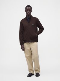 Homme portant une veste marron foncé zippée, un pantalon beige à jambes larges et des chaussures noires, debout devant un fond blanc uni.
