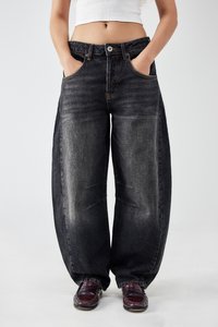 Persoon in losse, vervaagde zwarte jeans met de handen in de zakken en donkerbruine instappers, cropped wit shirt zichtbaar boven de taille. 