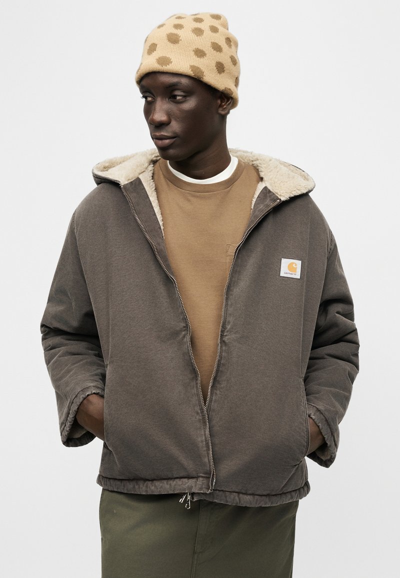 Mann mit beige gepunkteter Mütze, brauner Zip-Up-Jacke mit Fleecefutter, beige Hemd und olivgrüner Hose, die Hände in den Jackentaschen, schaut nach links.