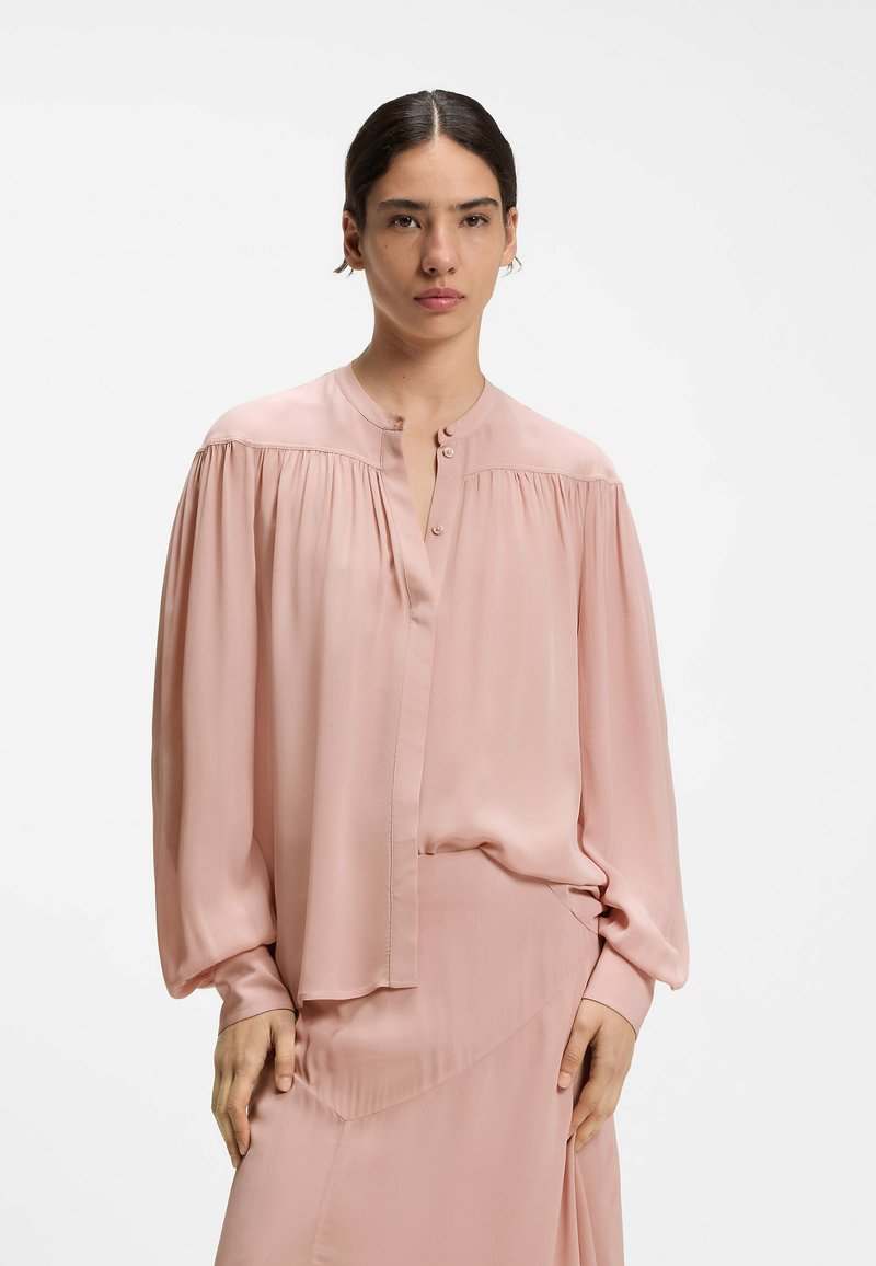 Femme aux cheveux foncés en chignon bas, portant un chemisier ample à manches longues rose pâle avec boutons et jupe assortie sur fond uni.