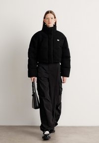 Svart cropped puffer-jakke med høy krage, teksturert fleece-materiale, frontlomme; parret med løse svarte cargo-bukser og en svart håndveske.