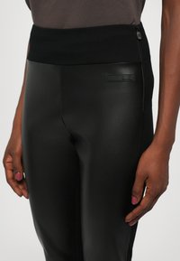 Black leggings with a high elastic waistband, featuring a smooth, textured finish and a side zip pocket. Includes a subtle brand logo.