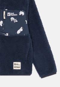 Pullover in pile blu con tasca a zip stampata sul petto con personaggi Moomin e un'etichetta con logo in basso a sinistra. Texture morbida.