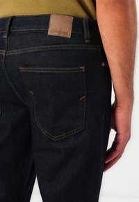 Dunkelblaue Denim-Jeans mit einem Lederpatch, das mit "Selected" beschriftet ist, kontrastiert durch orangefarbene Nähte. Subtiler Entwurf der Gesäßtasche.