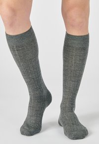 Gambe con calze grigie a coste fino al ginocchio, un piede piatto a terra, l'altro leggermente sollevato sulle punte contro uno sfondo semplice.