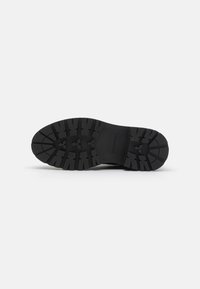 Semelle de chaussure en caoutchouc noir avec un motif de semelle profond, présentant une section lisse surélevée près du talon et une zone plate pour l'adhérence.
