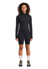 Svart atletisk topp med blixtlås fram, långa ärmar och logotyp; matchad med svarta cykelshorts. Buren med beige vandringsskor och vita strumpor.