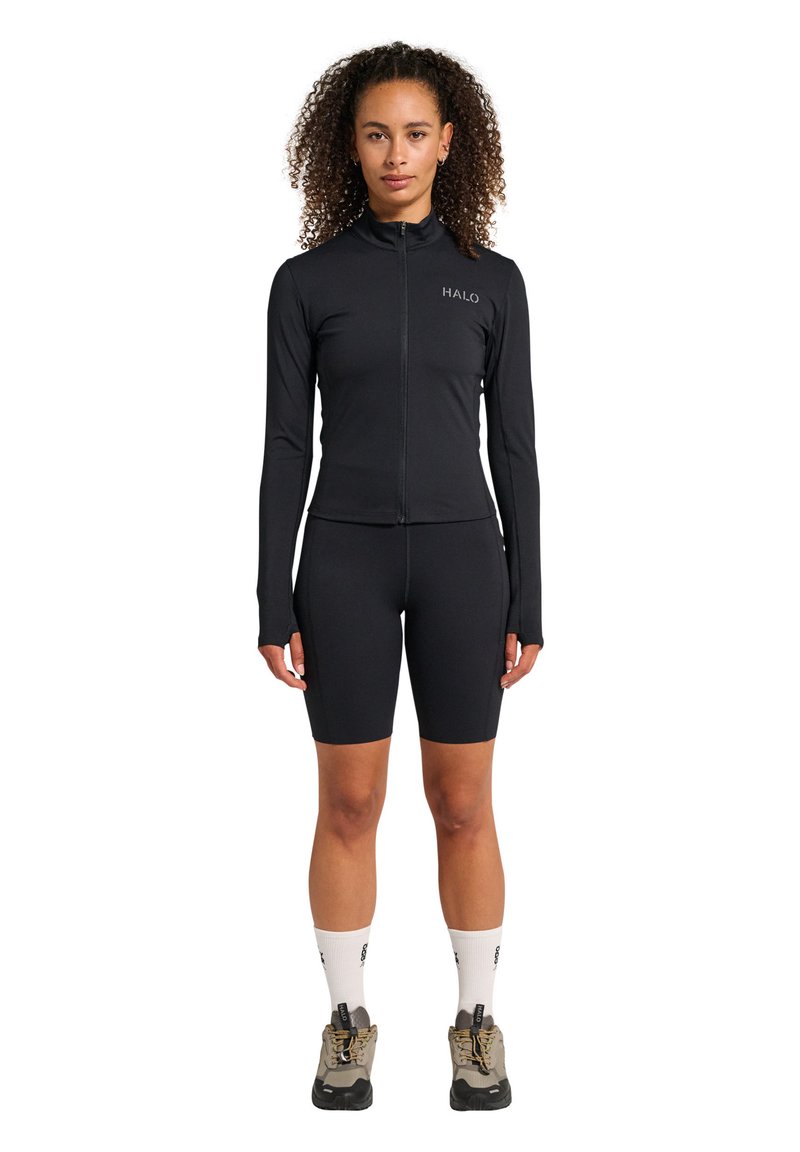 Svart atletisk topp med blixtlås fram, långa ärmar och logotyp; matchad med svarta cykelshorts. Buren med beige vandringsskor och vita strumpor.