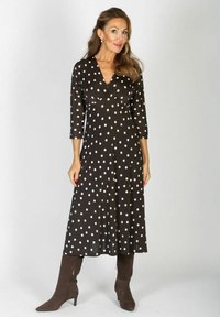 Kobieta z długimi włosami, ubrana w czarną sukienkę midi w białe kropki i brązowe buty na obcasie, stojąca na jednolitym tle.