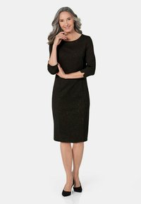 Schwarzes Kleid mit drei Viertelärmeln, taillierter Form, glitzernder Textur und knielangem Design, getragen mit schwarzen Absätzen. Einfacher runder Ausschnitt.