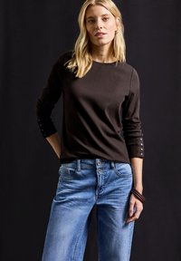 Braunes, geripptes Langarmoberteil mit Knopfakzenten an den Ärmeln, kombiniert mit hellblauen Jeans im Five-Pocket-Design und doppeltem Knopfverschluss.