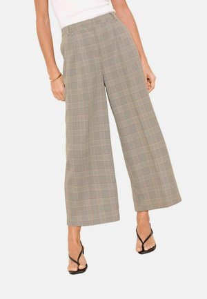 Femme portant un pantalon beige à carreaux taille haute à jambes larges, associé à des talons noirs à lanières, debout devant un fond uni.