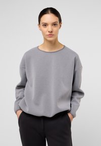 Felpa grigia chiaro con scollatura rotonda e spalle cadenti, caratterizzata da una texture morbida e una vestibilità rilassata. Abbinata a pantaloni scuri.