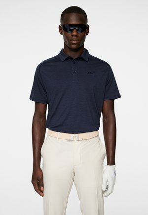 Navy polo shirt with a three-button placket and subtle texture, paired with beige trousers and a light glove. Black sunglasses complete the look.
