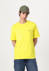 Žlté bavlnené tričko s krátkymi rukávmi a okrúhlym výstrihom, s malým zeleným logom Lacoste. Spárované s modrými džínsami a zelenou čiapkou.