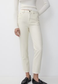 Pantalons en denim couleur crème avec une coupe slim, taille haute, design à cinq poches et détails de couture subtils, associés à des mocassins noirs.