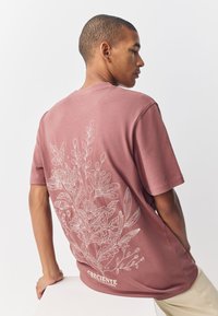 Maroon short-sleeve t-shirt featuring a white floral design on the back. Smooth texture with a relaxed fit. Brand logo at the bottom.