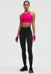 Leggings negras de cintura alta combinadas con un sujetador deportivo corto de color rosa brillante. La modelo lleva zapatillas deportivas negras y guantes sin dedos ajustados.