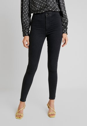 Kobieta w czarnych, obcisłych jeansach z wysokim stanem, czarno-białej bluzce w wzory i żółtych sandałkach na obcasie z paskami, stojąca na jednolitym tle.