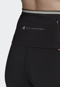 Czarne legginsy z wysokim stanem z srebrnym pasem, z kieszenią na zamek błyskawiczny i czerwonymi akcentami. Branding w białym tekście widoczny na pasie.