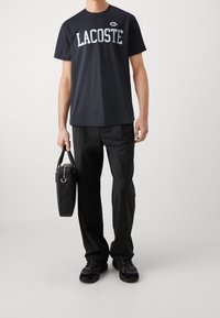 T-paita tummansinisestä Lacostesta, jossa on suuri logo, yhdistetty mustiin housuihin ja mustaan laukkuun. Kengät ovat mustat urheilutyyliset ja niissä on teksturoitu pinta.