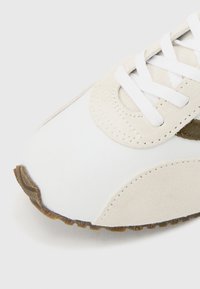 Baskets blanches et beiges vue de face avec des lacets blancs et une semelle texturée marron sur un fond clair.