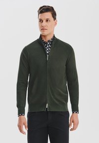 Ciemnozielony kardigan na zamek z miękkiego materiału, z kołnierzykiem i ściągaczami przy mankietach. Połączony z wzorzystą koszulą pod spodem.