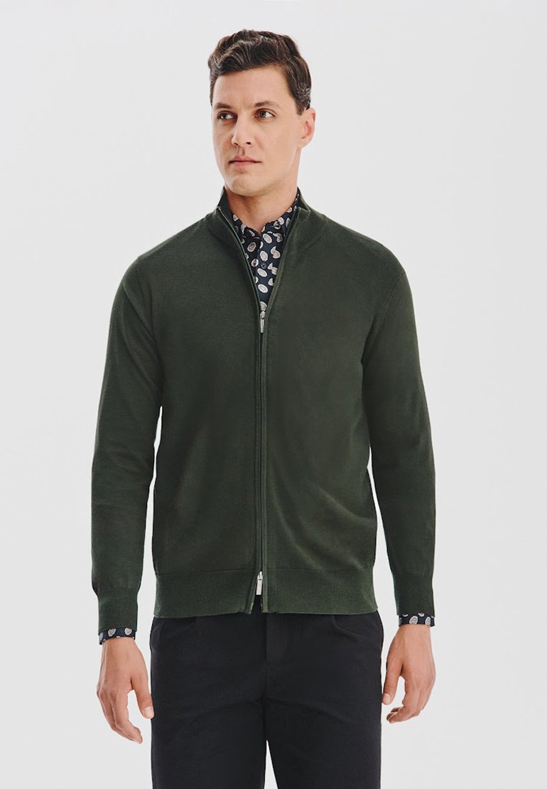 Ciemnozielony kardigan na zamek z miękkiego materiału, z kołnierzykiem i ściągaczami przy mankietach. Połączony z wzorzystą koszulą pod spodem.