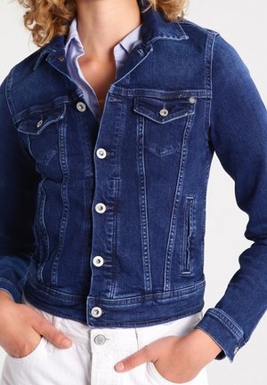 Personne portant une veste en jean bleu foncé cintrée par-dessus une chemise bleu clair, les mains dans les poches d'un pantalon blanc.