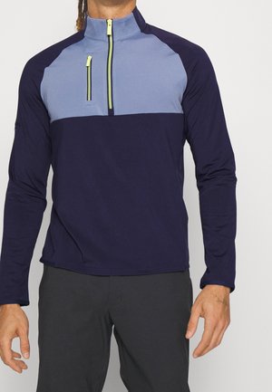 Mann trägt einen langärmligen, zweifarbigen navyblauen und hellblauen Half-Zip-Pullover mit neon-gelben Reißverschluss-Zippern, kombiniert mit schwarzen Hosen.