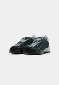 Sapatos de camurça verde escuro com detalhes em rede cinza, solas de borracha pretas e atacadores brancos em contraste. Apresenta uma alça na parte de trás.