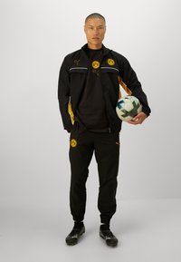 Sort atletisk træningsdragt med gule detaljer, der inkluderer en jakke med lynlås og bukser. Modellen holder en fodbold og har sorte fodboldstøvler på.