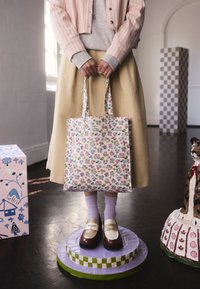 Osoba nosiaca béžovú sukňu a ružový pruhovaný kabát drží kvetinovú tašku, stojaca na fialovom podstavci v interiérovej umeleckej miestnosti.