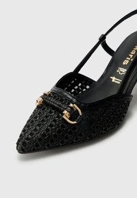 Zapato negro tipo slingback con punta afilada, textura tejida, detalle de metal dorado y una correa delgada. Cuero suave con un patrón intrincado.