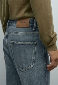 Vista posteriore di una persona che indossa jeans blu scoloriti con un'etichetta marrone Massimo Dutti e un maglione verde oliva.
