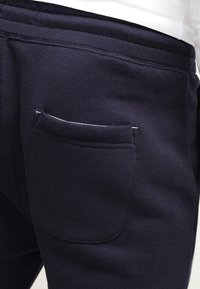 Mörkblå träningsbyxor i mjukt tyg, med en synlig bakficka med vit sömnad och elastisk midja.