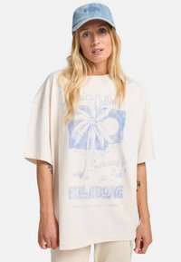 Lys beige oversized t-shirt lavet af bomuld, med et blåt grafik af et palmetræ og sol, med "BILLABONG" tekst nederst.