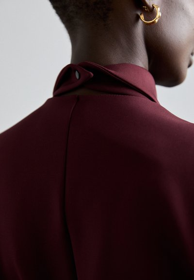 Rochie burgundă cu guler înalt și un buton decorativ la spate. Prezintă o textură netedă și linii curate. Cercei din aur în formă de cerc vizibili.