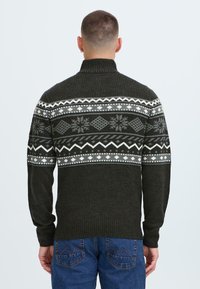 Mørkegrønn strikket genser med høy krage, med et geometrisk fair isle-mønster i grått og hvitt over den øverste delen.