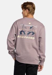 Purple sweatshirt with a crew neck, featuring a graphic of two figures carrying a surfboard, and the text "BILLABONG" in yellow.