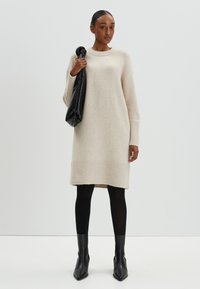 Robe pull en tricot beige avec des épaules tombantes, des poignets et un ourlet côtelés. Associée à des leggings noirs et des bottines pointues, tenant un sac noir.
