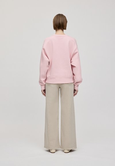 Felpa oversize rosa chiaro con maniche a pipistrello, indossata su pantaloni beige a gamba larga. Tessuto morbido con polsini e orlo a coste.