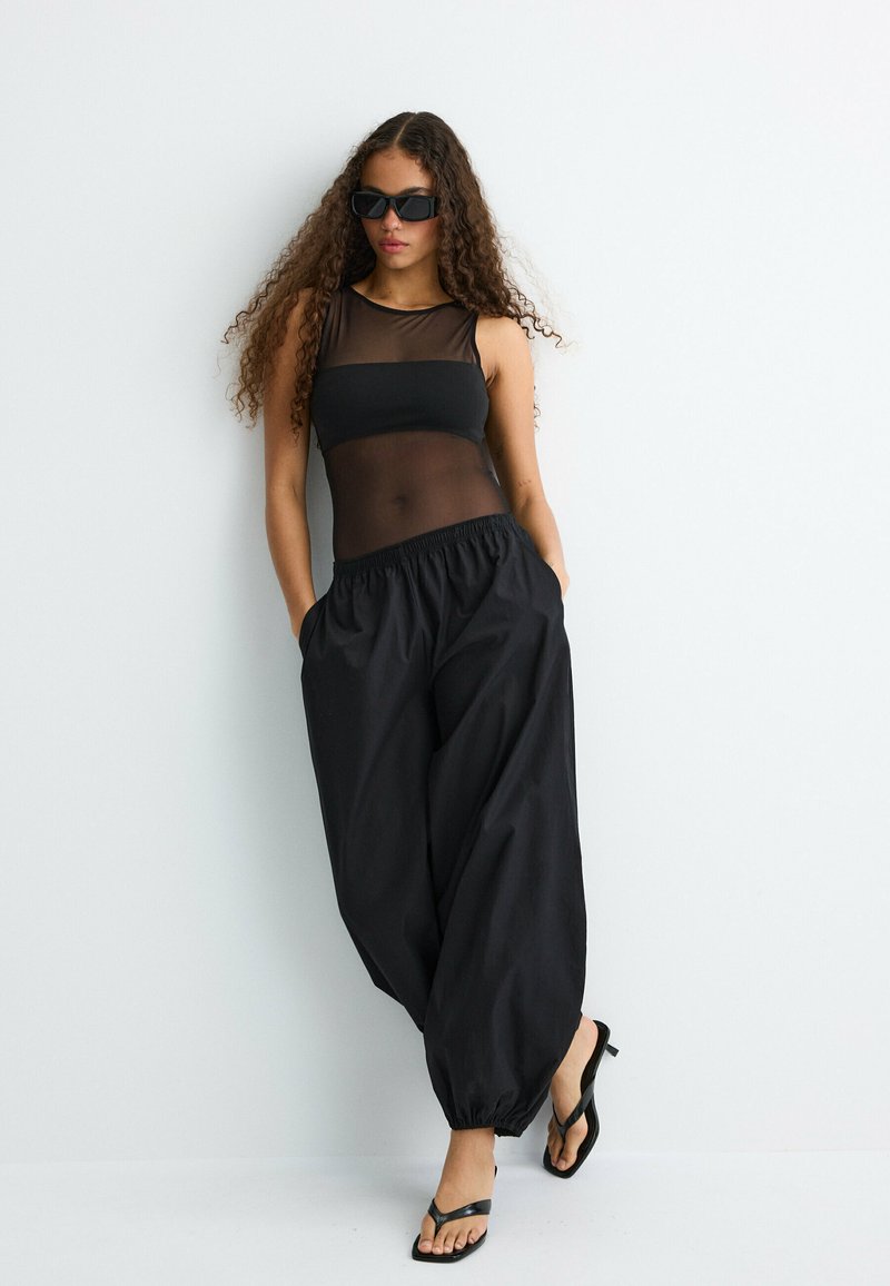 Femme aux longs cheveux bouclés porte un haut noir transparent sans manches, un pantalon noir ample, des sandales noires à talons et des lunettes de soleil foncées, posant devant un mur blanc.