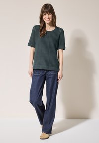 Maglione a maglia testurizzata verde scuro con maniche corte abbinato a jeans in denim scuri a gamba larga. Modella in piedi con le mani ai fianchi su uno sfondo chiaro.