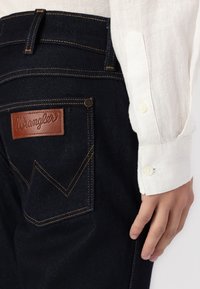 Tmavomodré rifľové džínsy s hnedou koženou náplasťou Wrangler, oranžové stehy, nosené s bielou košeľou s dlhým rukávom a rukou opretou vedľa nohy.