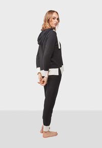 Sudadera negra y pantalones a juego con acentos en crema. Tela suave, corte relajado, con capucha y puños acanalados. El modelo posa de lado.