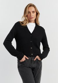 Cardigan nero realizzato in tessuto a maglia testurizzato, con scollo a V, chiusura con bottoni e polsini e orlo a coste. Abbinato a jeans grigio scuro.